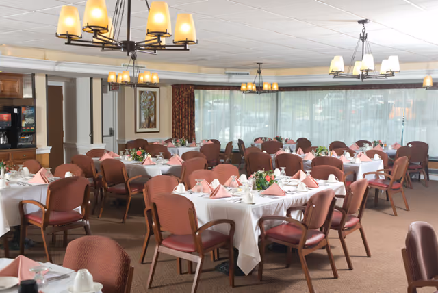 A dining room with multiple round tables covered with white tablecloths and set with pink folded napkins, cups, and plates. Each table has a floral centerpiece. The room has large windows with sheer curtains, brown chairs with armrests, and warm lighting from ceiling fixtures.