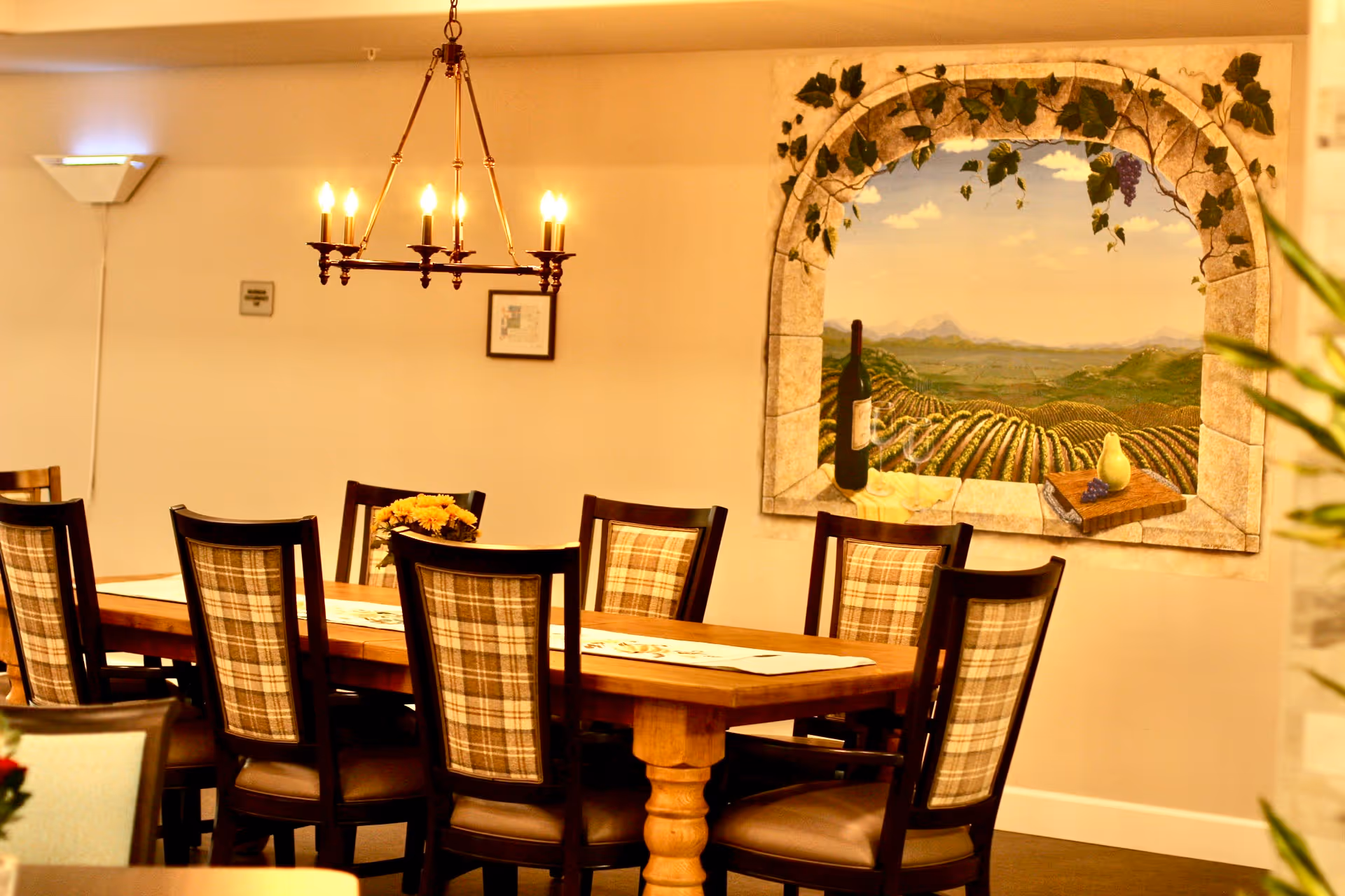 A dining room with a long wooden table surrounded by eight chairs with plaid backs. A chandelier with lit candles hangs above the table. On the wall, there is a large mural depicting a vineyard landscape framed by a stone arch with grapevines and a bottle of wine, a pear, and a cutting board with grapes in the foreground.
