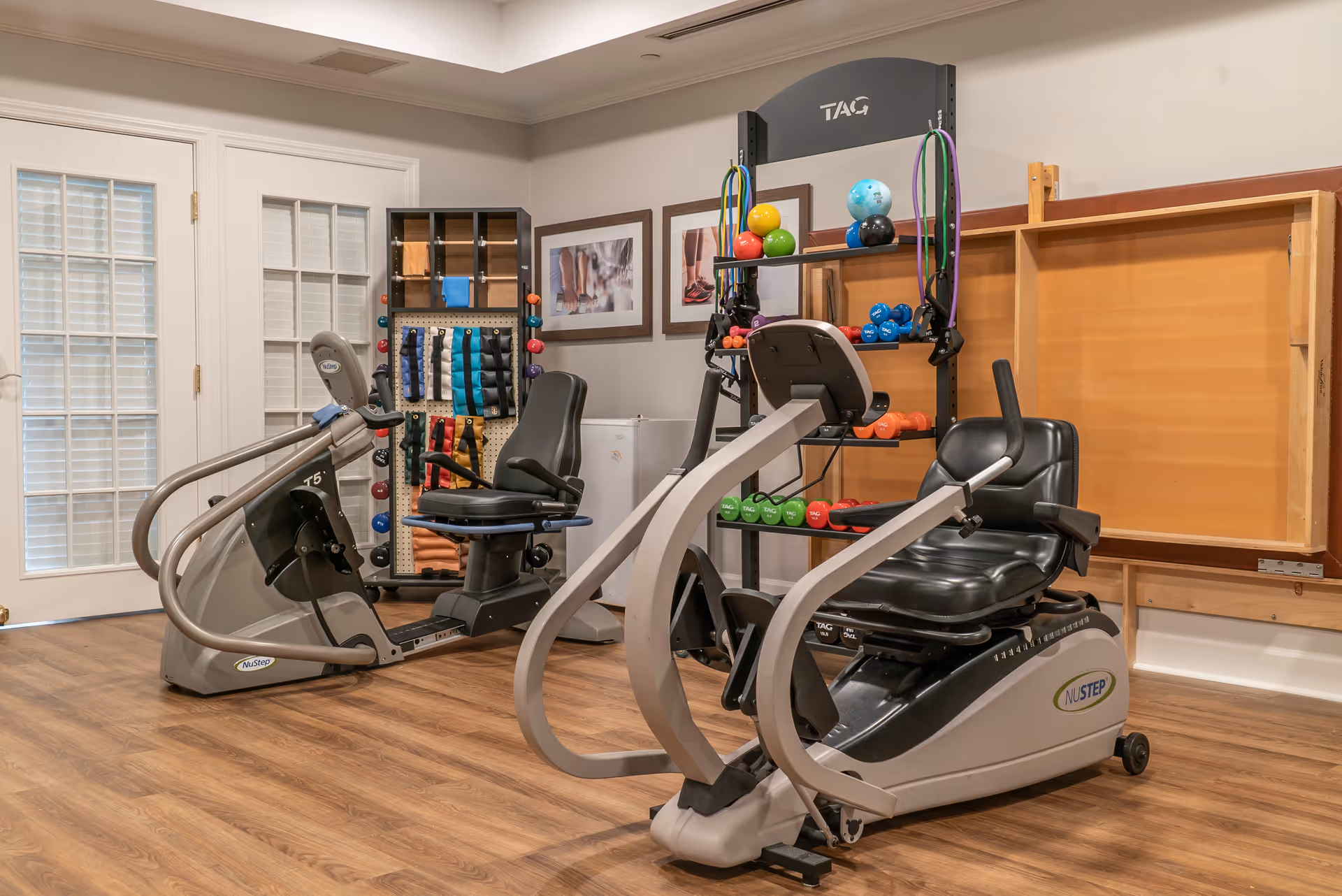 Small fitness room with two recumbent exercise machines, a rack of colorful dumbbells and resistance bands, and framed artwork on the wall.