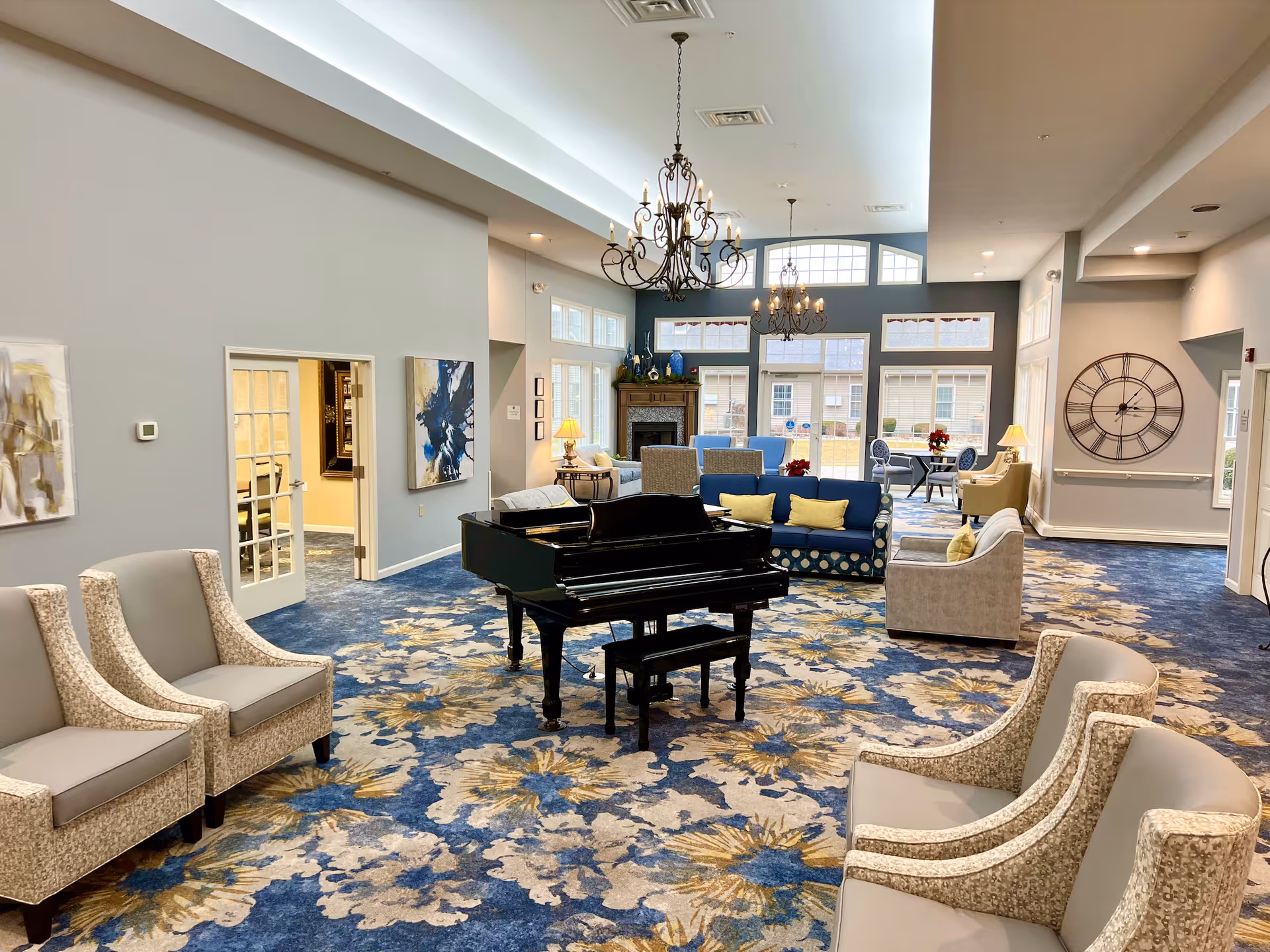 A spacious and well-lit living room area in an assisted living facility featuring a black grand piano in the center, multiple comfortable armchairs and sofas with cushions, a large floral patterned carpet in blue, beige, and gold tones, two ornate chandeliers hanging from the ceiling, large windows letting in natural light, a fireplace with decorative items on the mantel, and a large wall clock on the right wall.