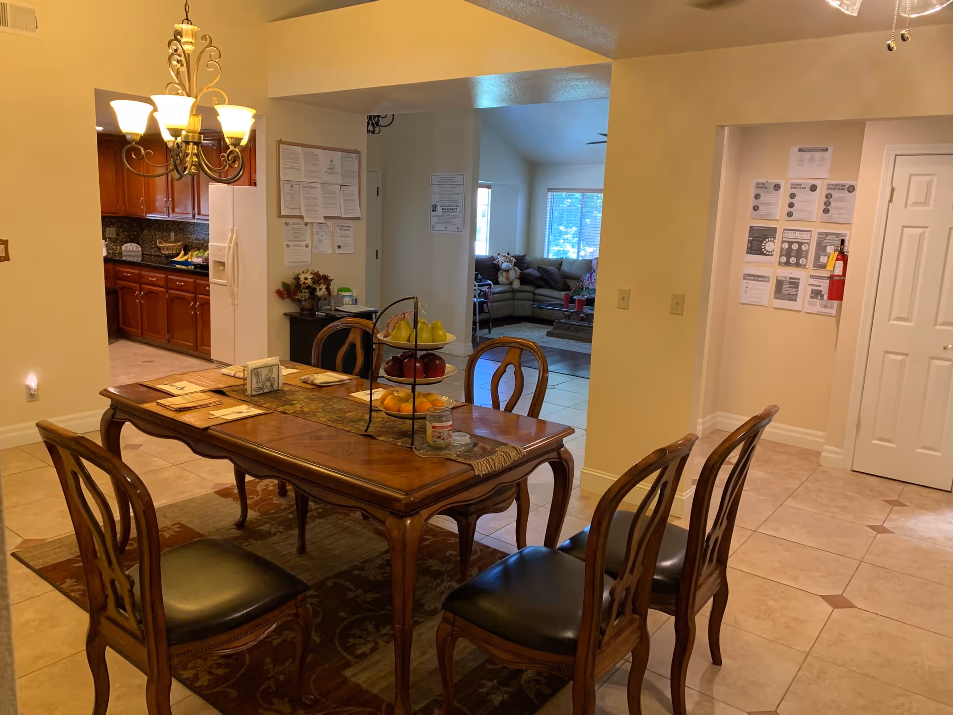 A dining area with a wooden table set for four with placemats and a tiered fruit stand in the center. The room has tile flooring and a chandelier hanging above the table. In the background, there is a kitchen with wooden cabinets and a refrigerator, and a living room with a sectional sofa and large windows letting in natural light. Walls have various notices and a fire extinguisher is mounted near a door.