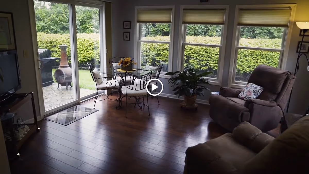 A cozy living room area with wooden flooring, featuring a glass-top table with four wrought iron chairs with cushions near large windows overlooking a green hedge. There is a brown recliner chair with a floral pillow, a potted plant, a floor lamp, and a sliding glass door leading to an outdoor patio with a chiminea and a barbecue grill.