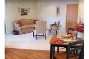 A cozy living area in Londonderry Village featuring a floral patterned sofa, a beige armchair, a small wooden side table with a lamp, and a dining table set for two with plates, glasses, and a vase of red flowers. The room has light-colored walls, a wooden floor near the dining area, and carpet in the seating area. There are two closed doors and framed artwork on the walls.