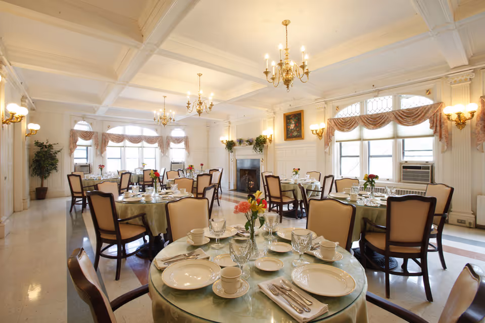 A bright and elegant dining room with round tables set with plates, cups, glasses, and silverware. Each table has a small floral centerpiece. The room features large windows with draped curtains, chandeliers hanging from a coffered ceiling, wall sconces, and a fireplace with a painting above it. Several upholstered chairs surround each table.