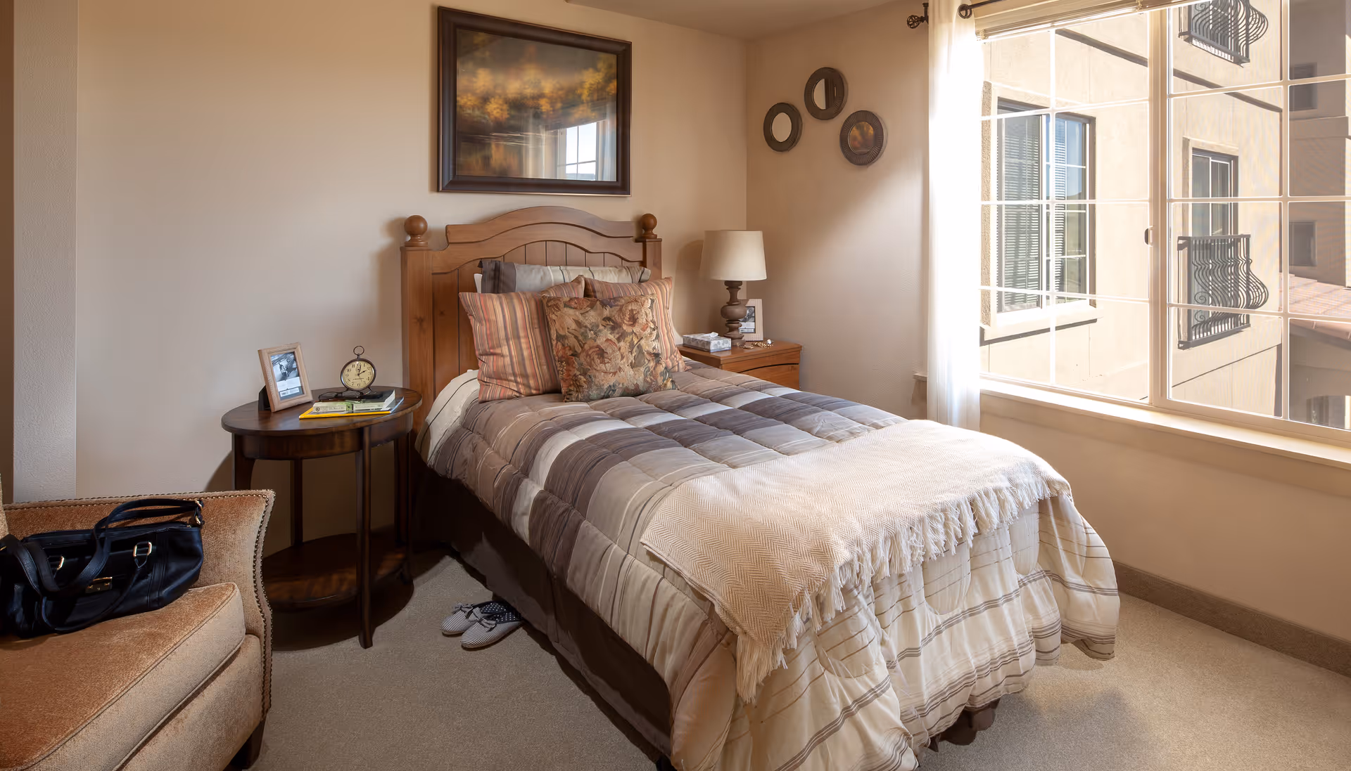 A cozy senior living bedroom with a single bed made with a striped comforter and decorative pillows. There is a wooden headboard, a nightstand with a lamp, picture frame, and books. A round side table holds a clock, a photo frame, and a book. A cushioned armchair with a black handbag is placed near the bed. The room has a large window with sheer curtains letting in natural light, and the walls are decorated with framed artwork and circular wall hangings.