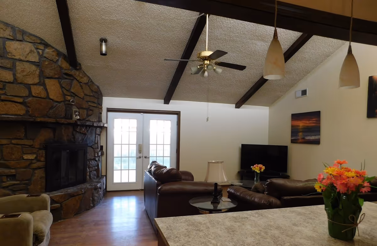 A cozy living room with a stone fireplace, brown leather furniture, and large windows.