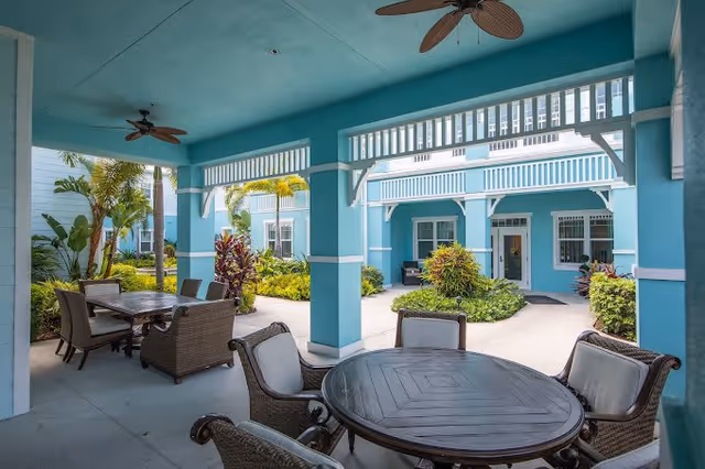 Covered outdoor seating area with round and rectangular tables surrounded by cushioned wicker chairs, blue painted columns and walls, tropical plants, and a view of the building exterior with windows and doors.