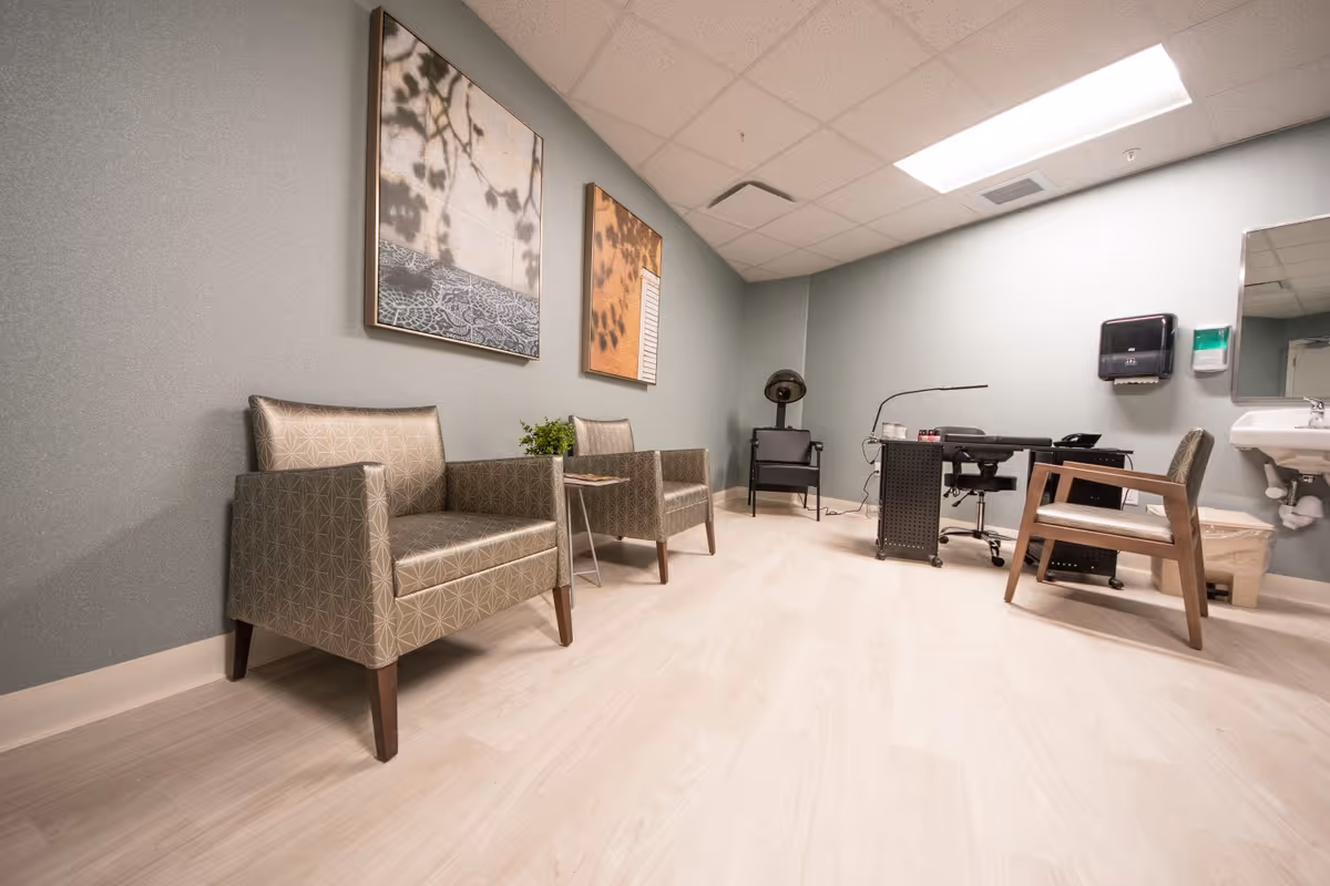 A clean salon/waiting room with two patterned armchairs, a manicure table and salon chair, a hooded hair dryer, and a wall-mounted sink.
