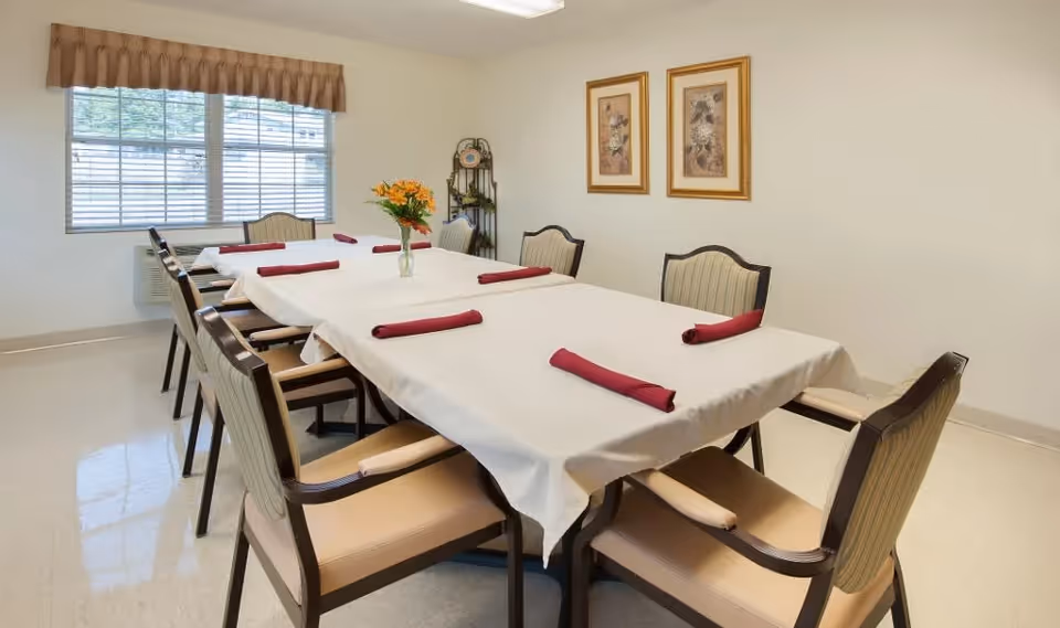 A dining room with a rectangular table covered with a white tablecloth and eight chairs around it. Each place setting has a rolled red napkin. A vase with yellow flowers is placed in the center of the table. The room has a window with blinds and a valance, two framed floral pictures on the wall, and a small decorative shelf in the corner.