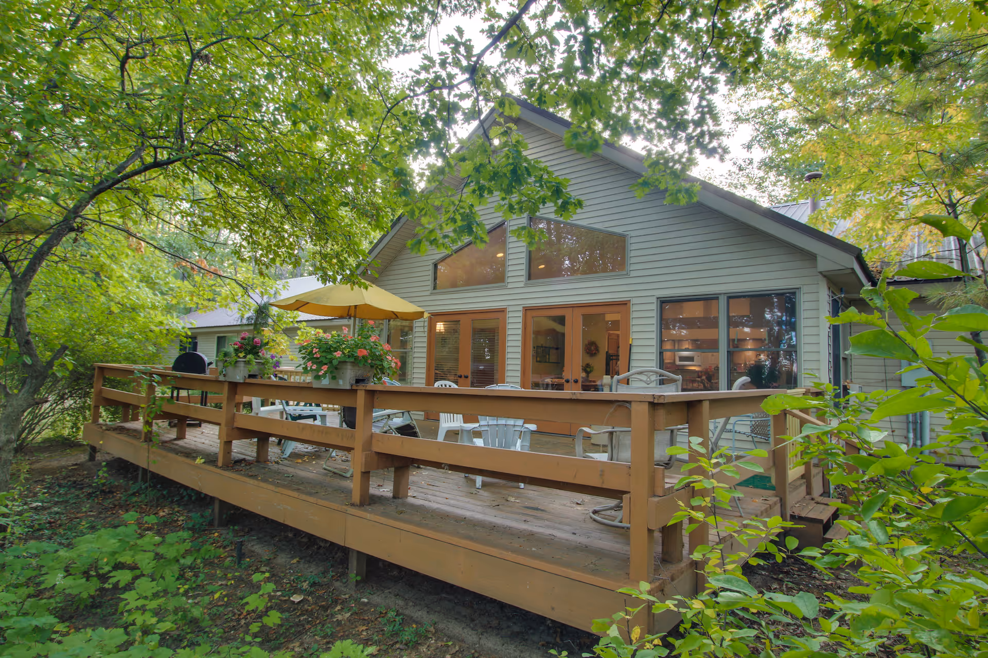 Exterior view of a single-story building with a wooden deck surrounded by lush green trees and plants. The deck has outdoor furniture including chairs, a table with a yellow umbrella, and flower pots. Large windows and glass doors provide a view into the interior of the building.