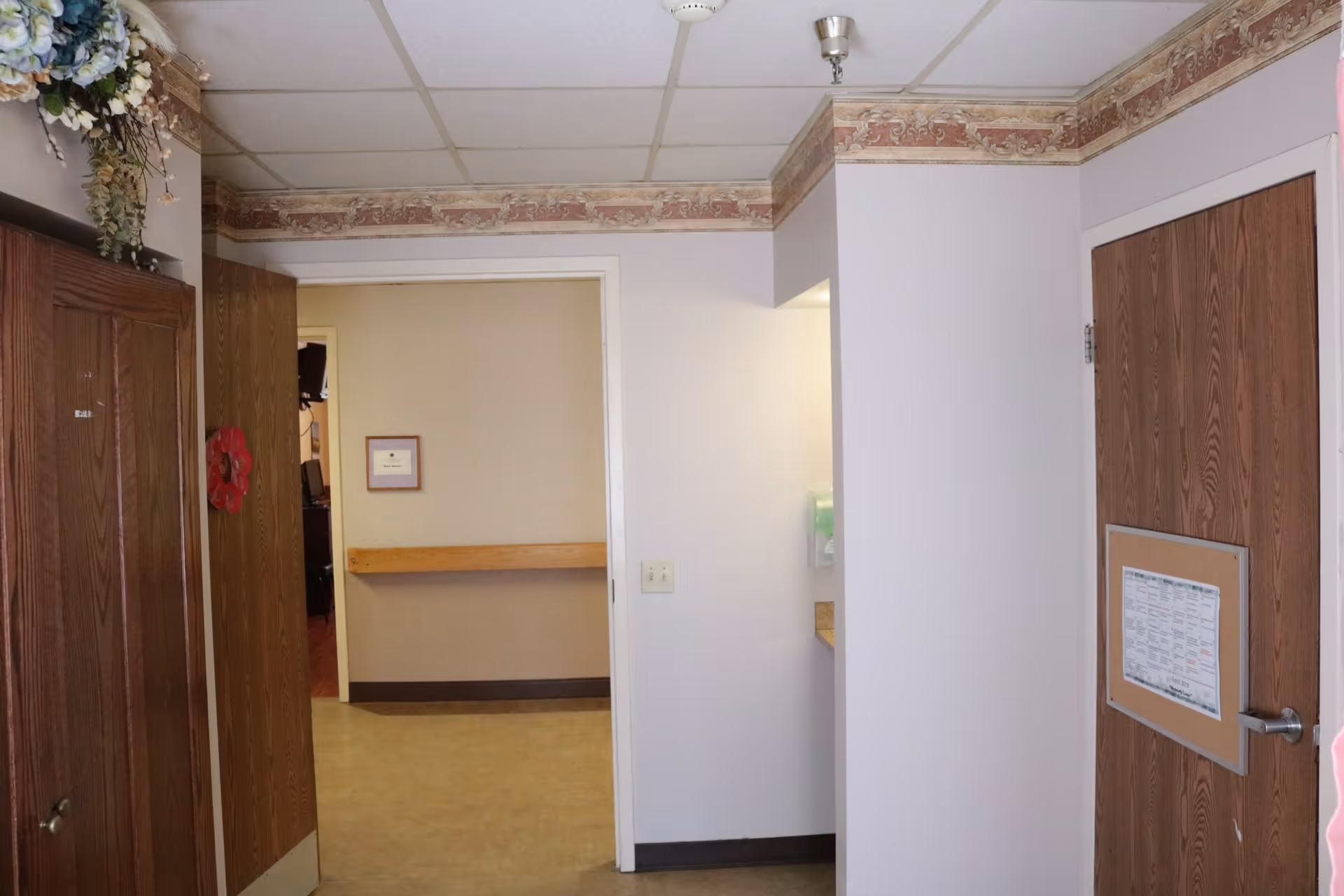 Interior hallway of a senior living facility with wood-paneled doors, a beige wall with a framed certificate, a wooden handrail, and floral wallpaper border near the ceiling. There is a small alcove with a mounted dispenser on the right side.