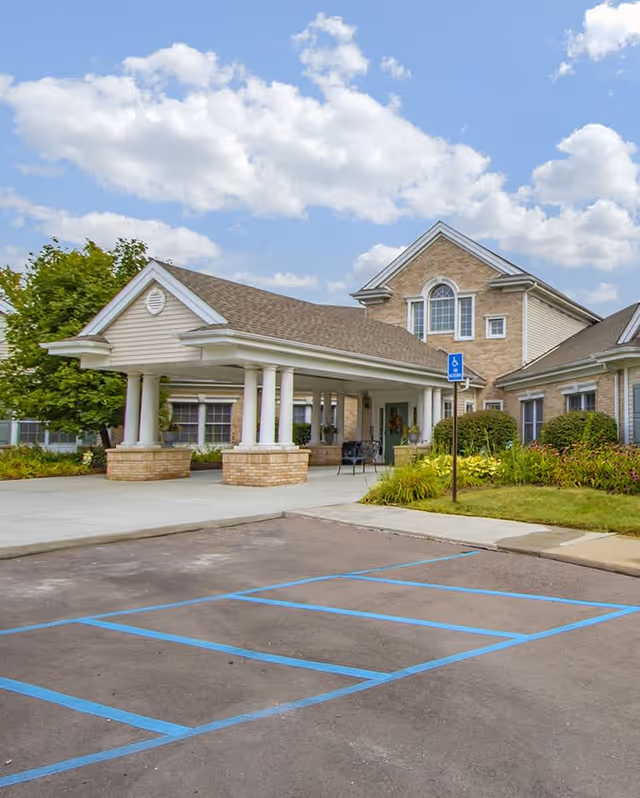 Exterior view of a senior living facility with a covered entrance supported by white columns, a brick and siding facade, a handicapped parking space in the foreground, and a partly cloudy sky.