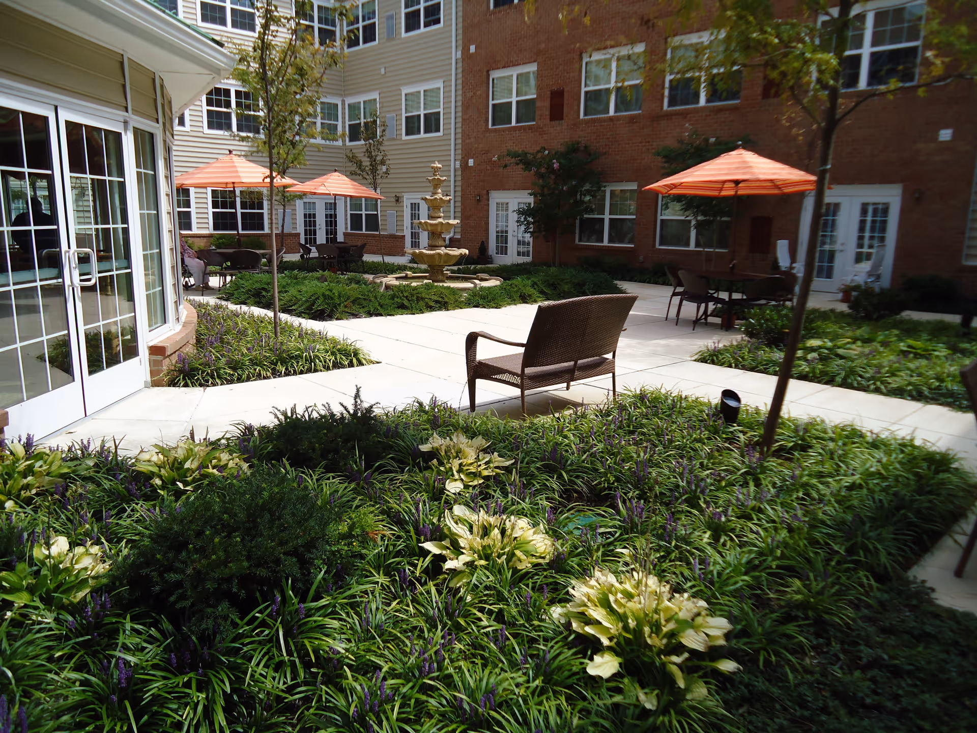 Outdoor courtyard area of a senior living facility with green plants, small trees, a central multi-tiered fountain, patio tables with orange umbrellas, and a bench. The courtyard is surrounded by a multi-story building with windows and doors.