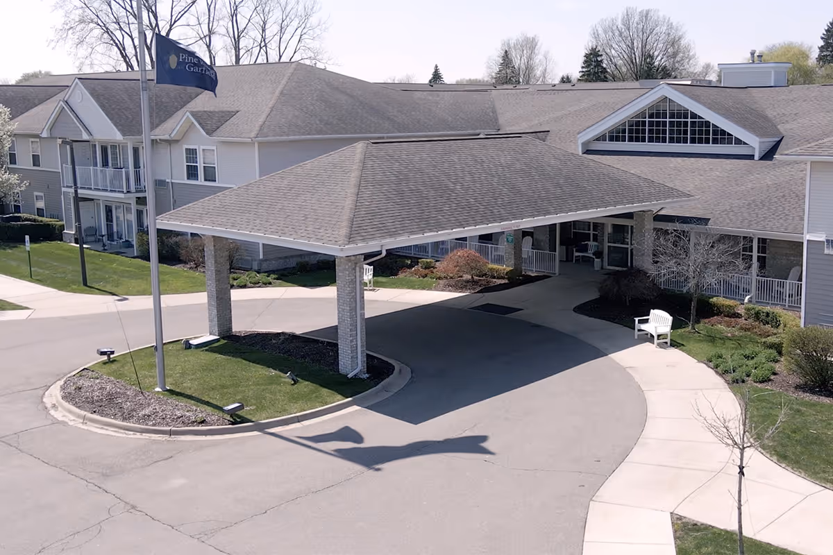 Exterior view of Pine Ridge of Garfield senior living facility showing the main entrance with a covered drop-off area, flagpole with a flag, landscaped greenery, and a paved driveway.