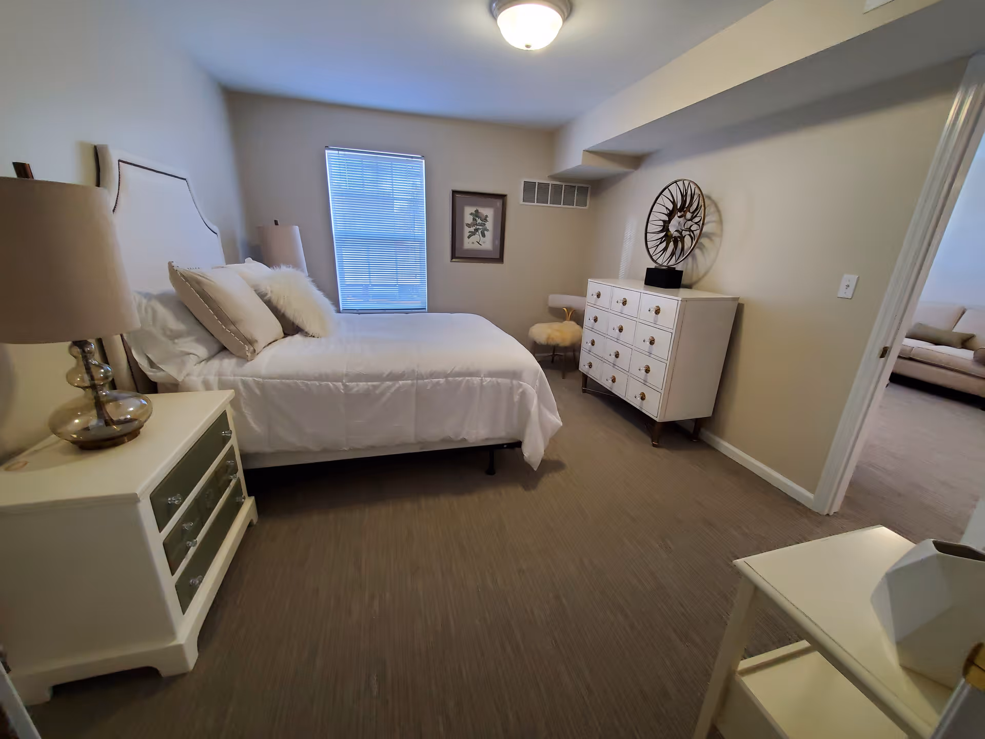 A bedroom with a neatly made bed featuring white bedding and multiple pillows. There is a bedside table with a lamp on the left side of the bed. A window with blinds is centered on the far wall, and a framed picture hangs to the right of the window. On the right side of the room, there is a white dresser with multiple drawers and a decorative sculpture on top. A small cushioned chair with a furry seat is next to the dresser. The room has beige walls and carpeted flooring. An open doorway leads to another room with a sofa visible.