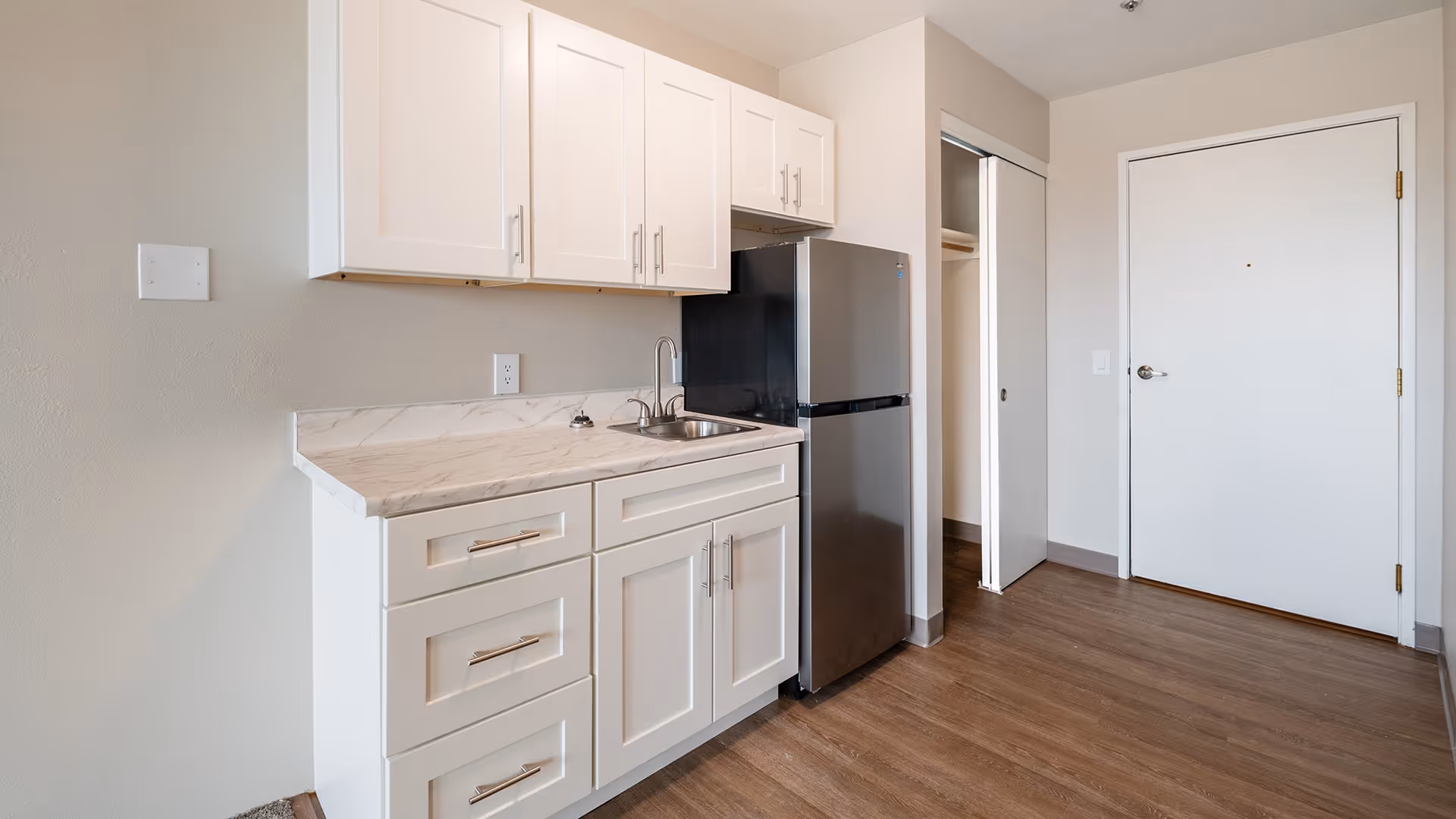 A small kitchen area with white cabinets, a marble countertop, a stainless steel sink, and a compact stainless steel refrigerator. The floor is wood laminate, and there is a white door and a closet with sliding doors in the background.