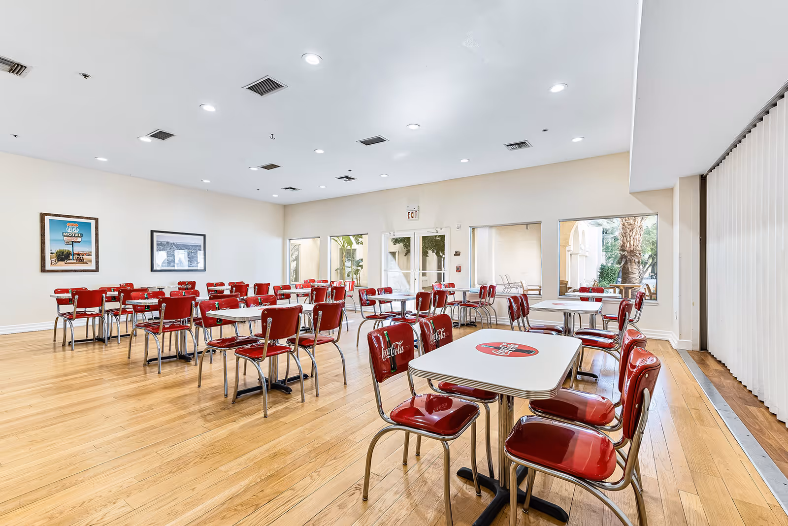 Bright communal dining room with rows of retro red Coca-Cola chairs and white tables on a hardwood floor.