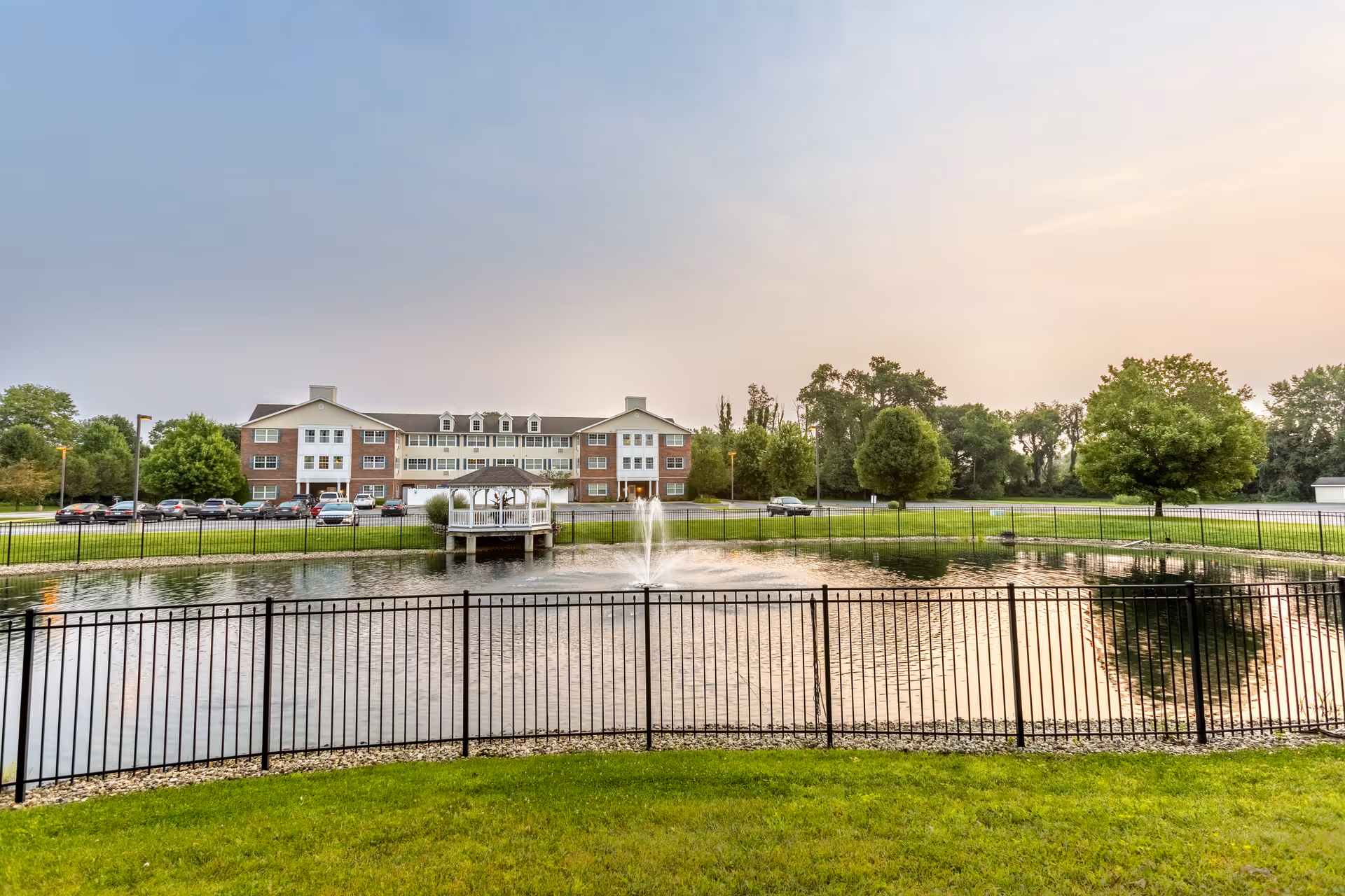 A serene outdoor scene at StoryPoint Granger featuring a large pond with a central water fountain, surrounded by a black metal fence. In the background, there is a three-story brick and white building with multiple windows and a gazebo extending over the pond. The area is landscaped with green grass and trees under a partly cloudy sky during sunset.