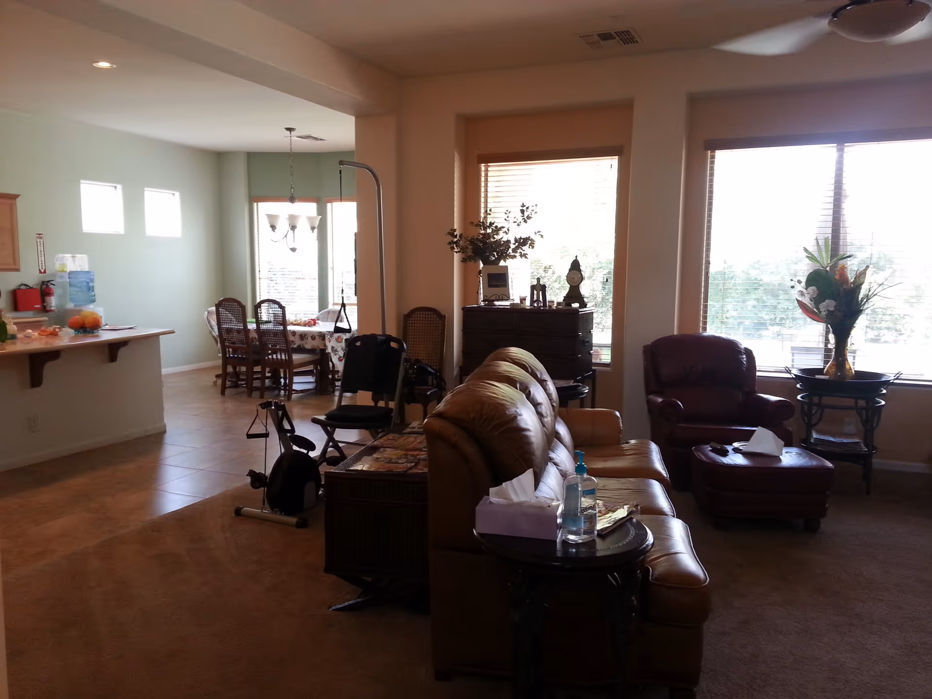 Interior view of a senior living facility showing a living room area with a brown leather sofa, a side table with tissues and hand sanitizer, a maroon armchair with an ottoman, and a decorative table with a flower vase near large windows with blinds. In the background, there is a dining area with a table and chairs, a kitchen counter with fruit and a water dispenser, and exercise equipment.