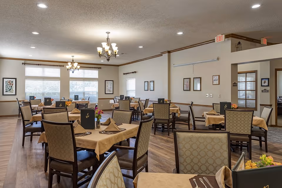 A well-lit dining room with multiple tables covered in beige tablecloths, each set with folded napkins, menus, and silverware. The room has wooden flooring, beige walls with framed artwork, and large windows letting in natural light. Ceiling chandeliers provide additional lighting.