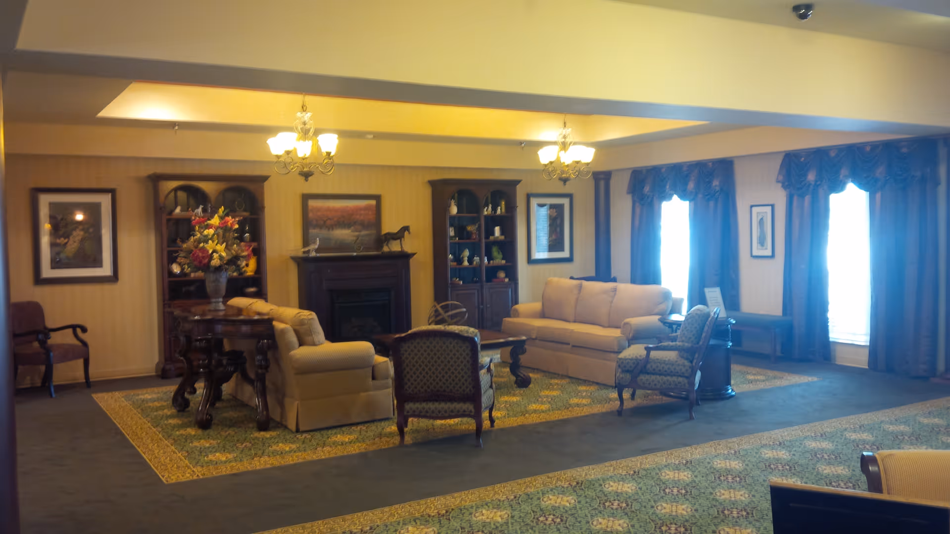 A spacious living room area with beige sofas, patterned armchairs, a wooden coffee table, and a fireplace flanked by two wooden bookshelves. The room has warm lighting from chandeliers, framed artwork on the walls, large windows with blue curtains, and a decorative rug covering part of the carpeted floor.
