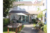 Outdoor courtyard area of a senior living facility with a paved walkway, green grass, plants, and two green patio umbrellas shading tables and chairs. The building has light-colored siding and multiple windows.
