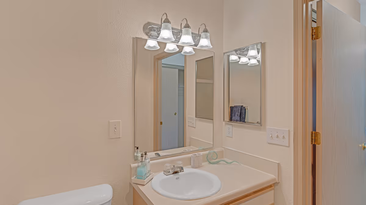 A bathroom vanity area with a white sink, countertop, and a large mirror above it. There is a four-light fixture mounted above the mirror. A soap dispenser and a small decorative item are on the countertop. A partially open door and a towel rack with a towel are visible in the reflection of the mirror.