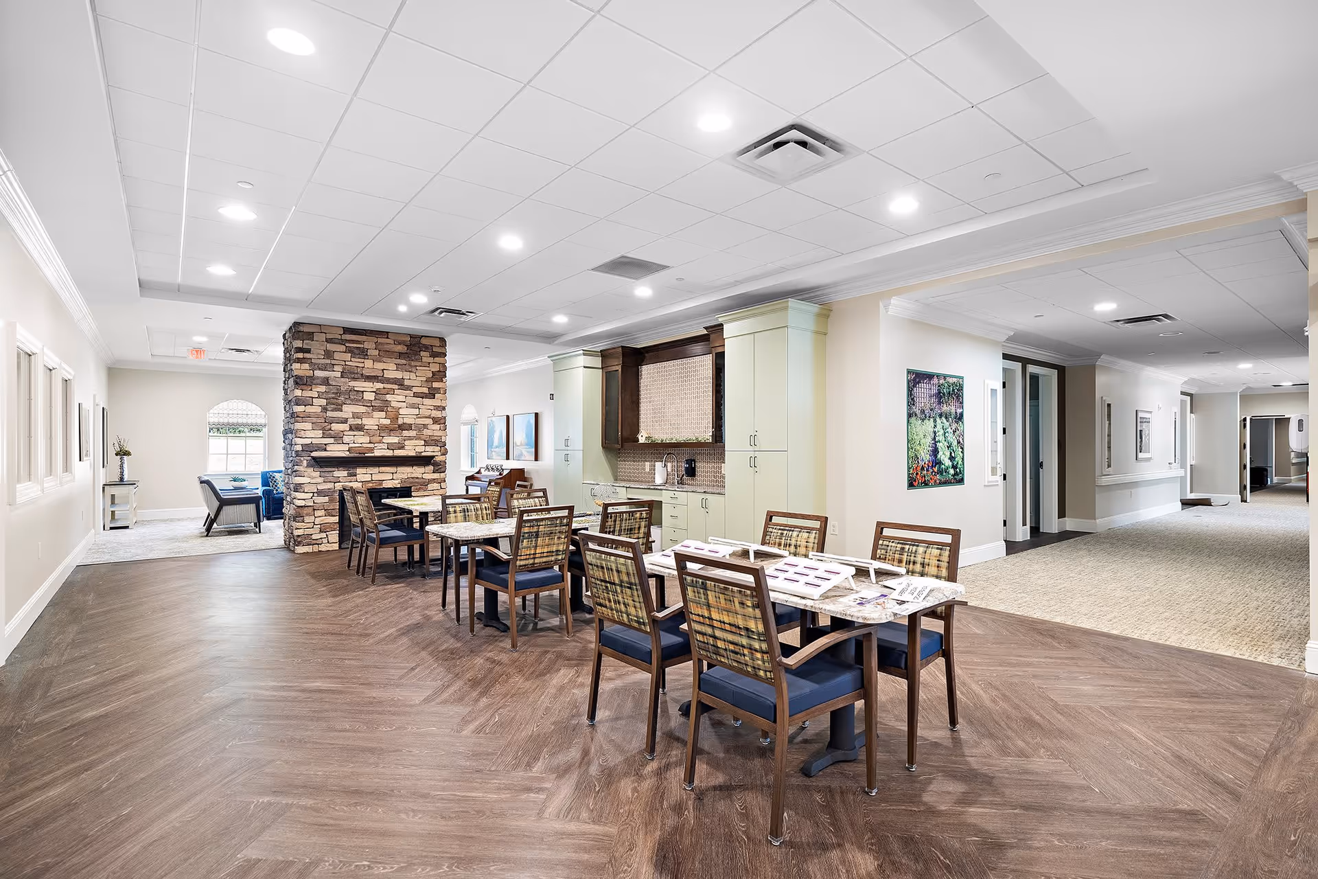Interior view of a senior living facility common area with a dining section featuring tables and chairs, a stone fireplace, and a kitchenette with cabinets. The space is well-lit with recessed ceiling lights and has a hallway leading to other rooms.