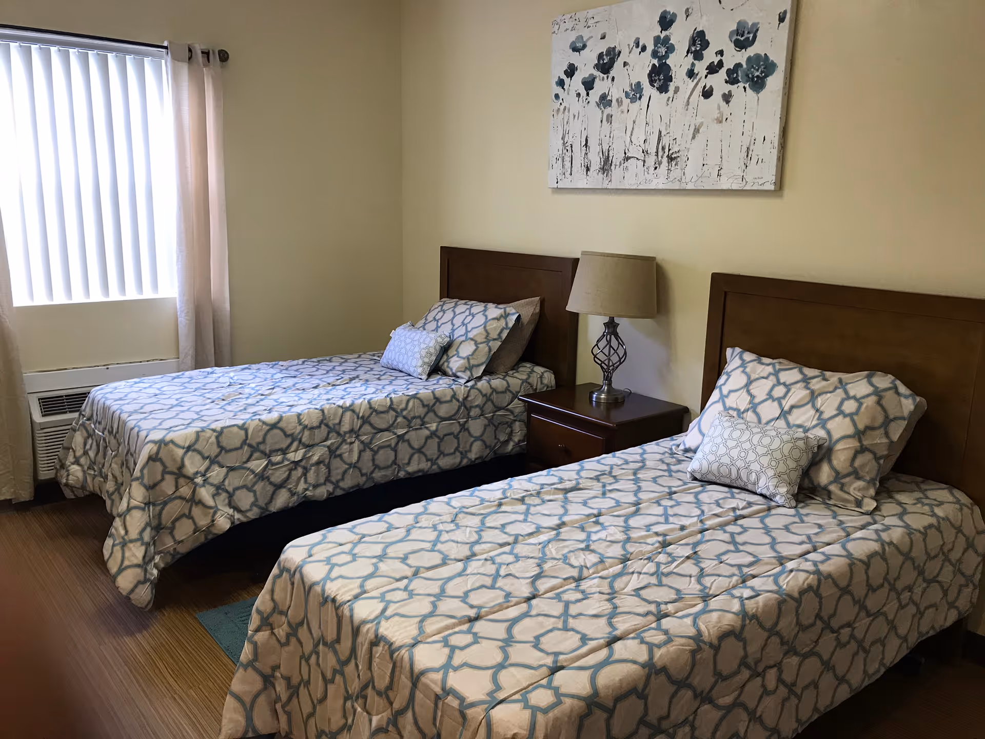 A simple bedroom with two twin beds featuring matching patterned bedding, a nightstand with a lamp, and a window with vertical blinds.
