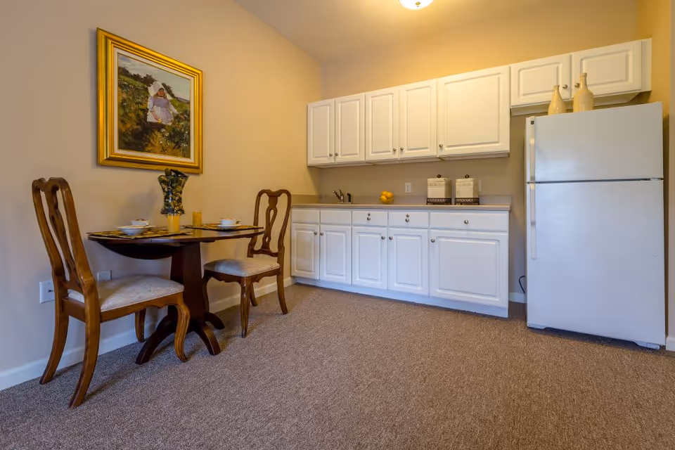 A small kitchen area with white cabinets, a white refrigerator, and a countertop with a sink. Next to the kitchen is a round wooden dining table set for two with two wooden chairs. A framed painting hangs on the beige wall above the dining table.