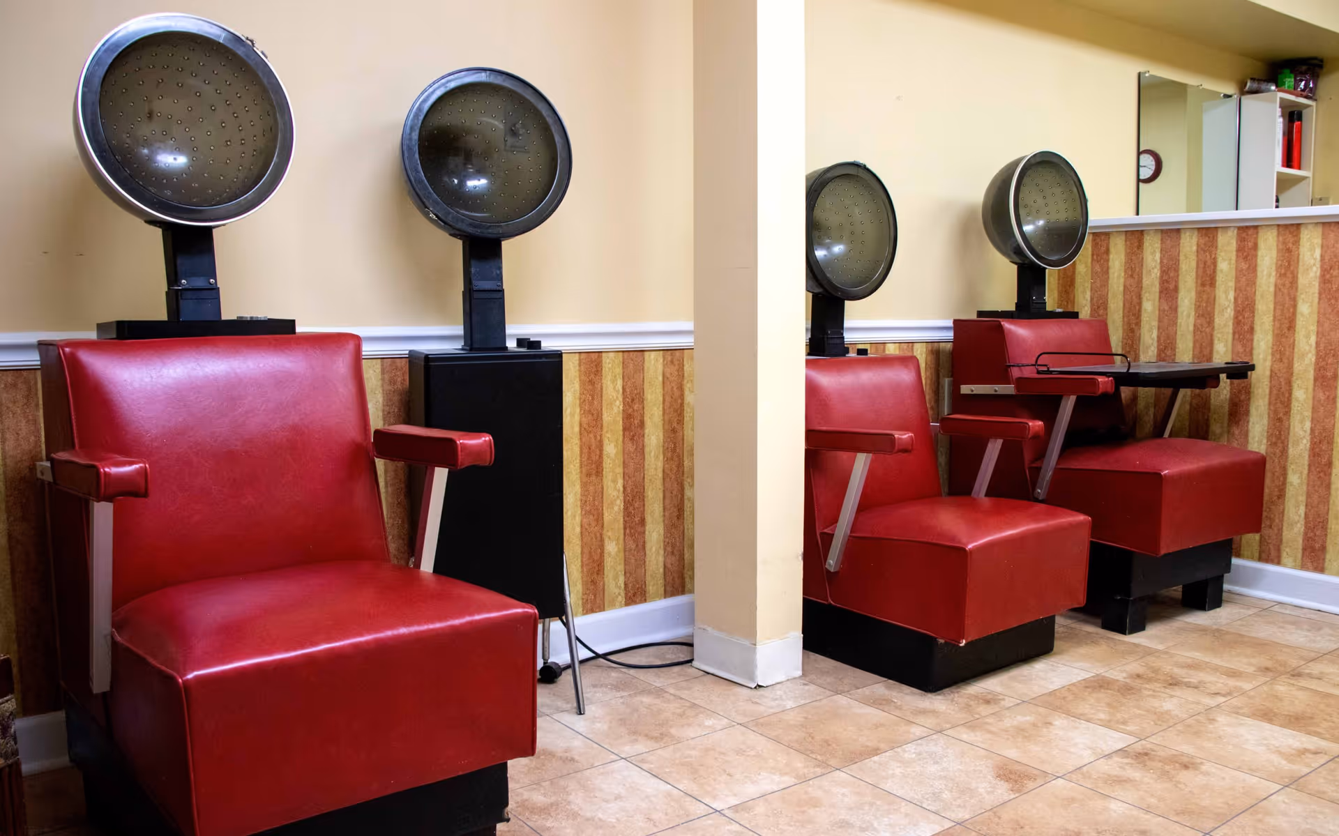 Salon area with three red vinyl drying chairs and overhead hair dryers against a striped wall.