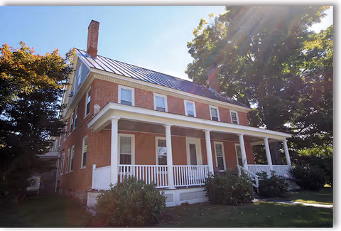 A large two-story brick house with a white porch railing and columns, surrounded by green bushes and trees under a clear blue sky with sunlight filtering through the leaves.