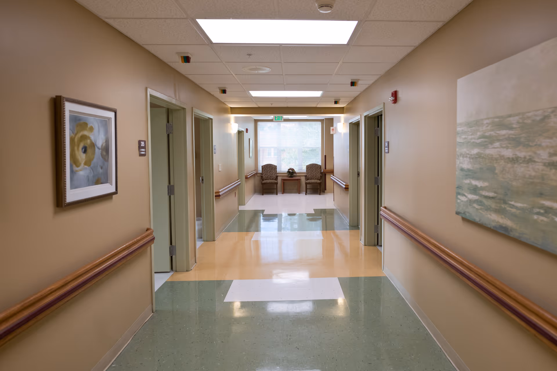 A clean, well-lit hallway in a healthcare facility with beige walls and a polished floor featuring green, yellow, and white sections. The hallway has handrails on both sides and several open doorways. At the end of the hallway, there are two chairs and a small table in front of a large window letting in natural light. Artwork is hung on the walls.