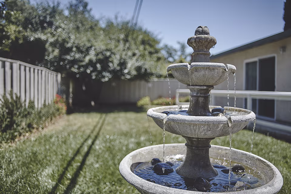 A three-tier stone water fountain in a grassy backyard area with a wooden fence and trees in the background, next to a building with sliding glass doors.