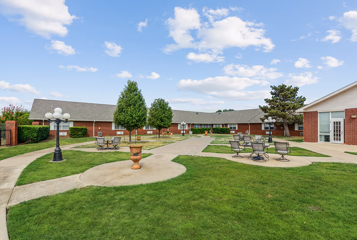 Courtyard of a single-story brick senior living facility with walkways, patio tables, lamp posts, and grassy lawns under a blue sky.