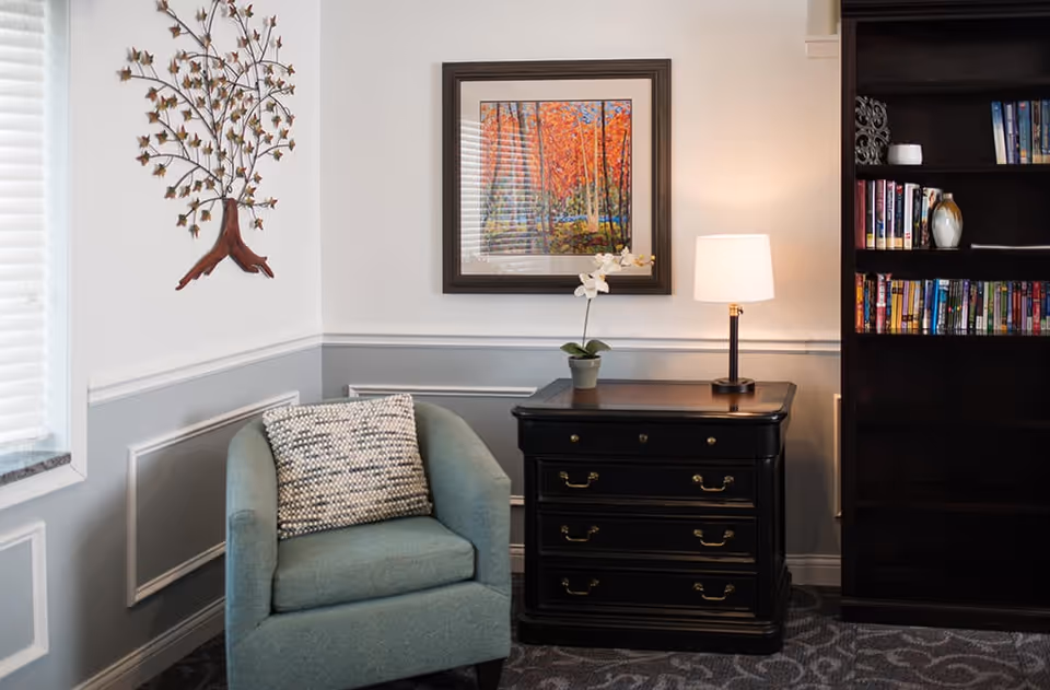 Cozy reading nook with a teal armchair and pillow, black side table with a lamp and potted orchid, framed artwork on the wall, and a bookshelf.