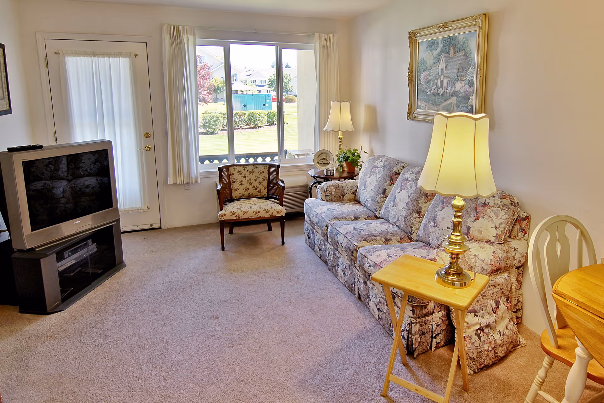 A cozy living room with a floral patterned sofa, a wooden side table with a lamp, a floral upholstered chair, a television on a stand, and a window with white curtains showing a view of a green lawn outside.