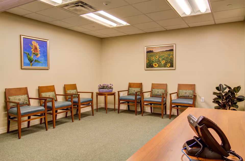 A waiting room with a row of wooden chairs along beige walls, two framed sunflower paintings, a small table with flowers, and a desk with a telephone in the foreground.
