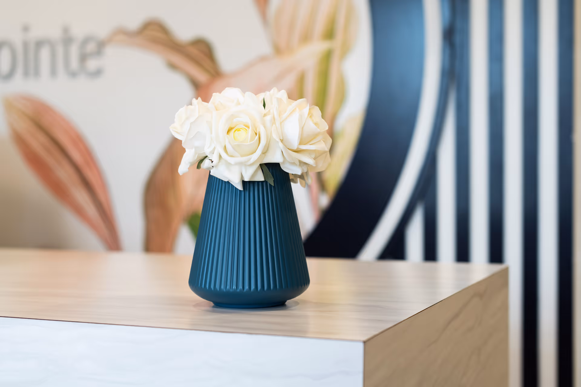 A teal ribbed vase holding white roses sits on a light wood reception desk with a blurred decorative background.