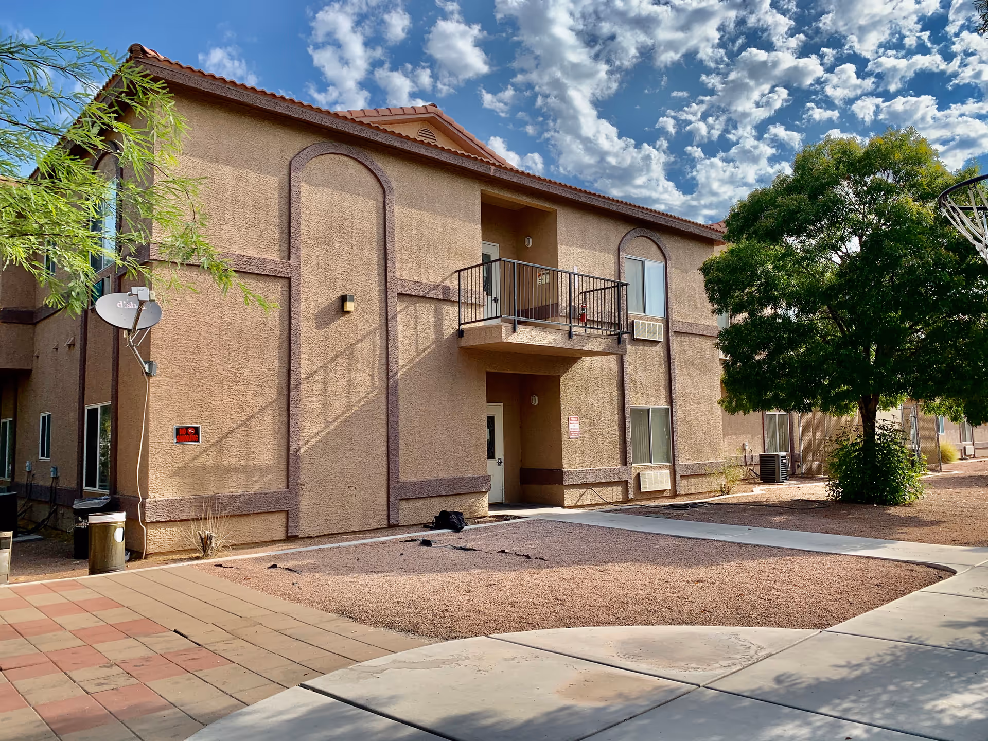 Exterior view of a two-story building with a balcony, several windows, and a tree on the right side under a partly cloudy sky.