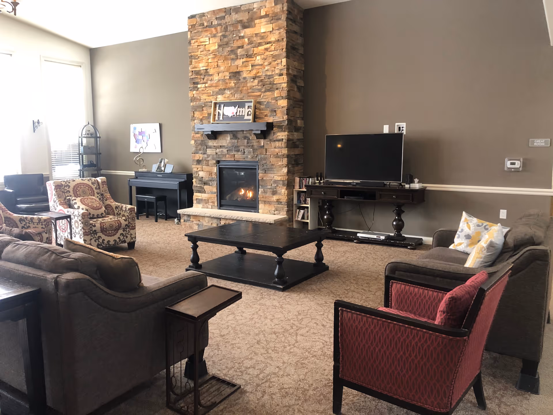 A cozy living room with a stone fireplace in the center, a flat-screen TV on a dark wooden stand to the right, and various seating options including a gray sofa, a patterned armchair, a red armchair, and another sofa with decorative pillows. There is a black coffee table in the middle and a piano against the far wall. The walls are painted a warm beige color.