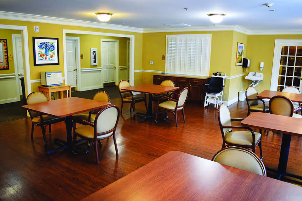 A dining room with multiple wooden tables and cushioned chairs arranged neatly on a polished wooden floor. The walls are painted yellow with white trim, decorated with framed artwork. There is a small sink and a chair with armrests in the corner, and several doors and windows with white shutters are visible.
