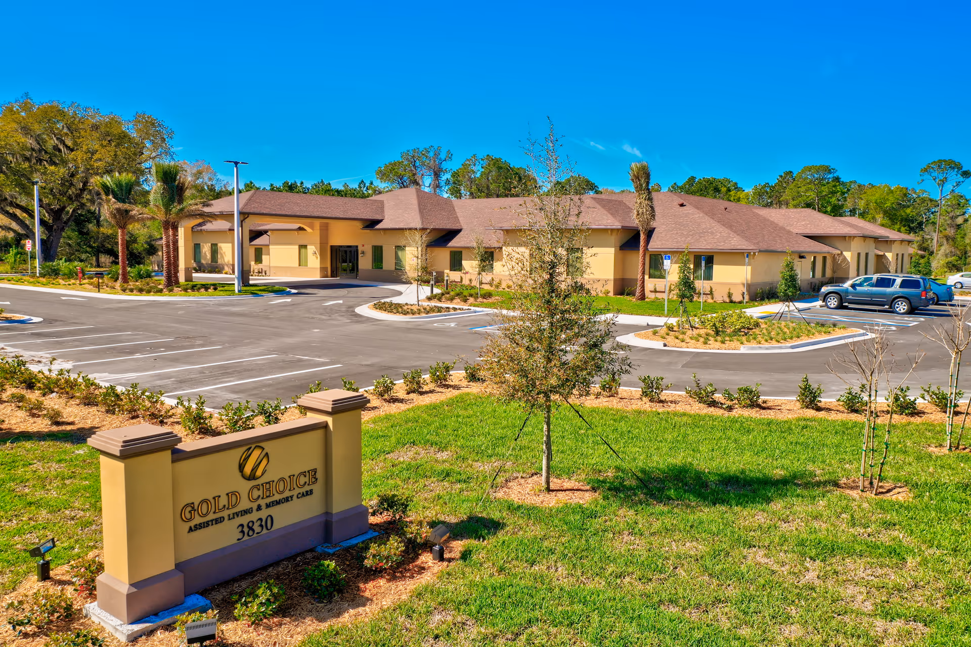 Exterior view of Gold Choice Palm Coast Assisted Living & Memory Care building with a clear blue sky, landscaped greenery, a parking lot with several cars, and a sign displaying the facility name and address 3830.