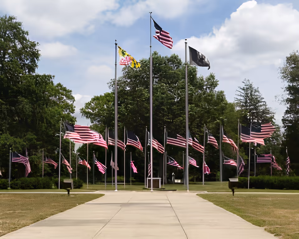 A paved walkway leading to a circle of tall flagpoles displaying multiple American and other flags on a grassy, tree-lined lawn.