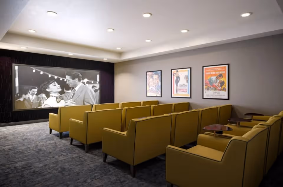 A small movie theater room with rows of yellow armchairs facing a large screen showing a black and white movie. The walls are decorated with three framed vintage movie posters. The room has recessed ceiling lights and a carpeted floor.