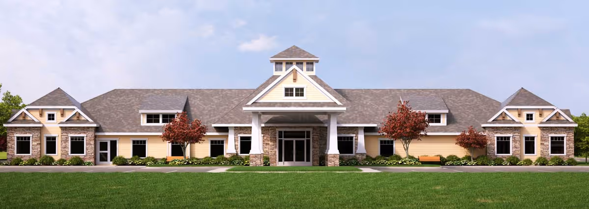 Front exterior view of a single-story senior living facility building with a central entrance covered by a porch supported by white columns. The building has a beige and stone facade with multiple windows and a gray shingled roof. There are small trees and shrubs planted along the front, and a well-maintained green lawn in the foreground under a partly cloudy sky.