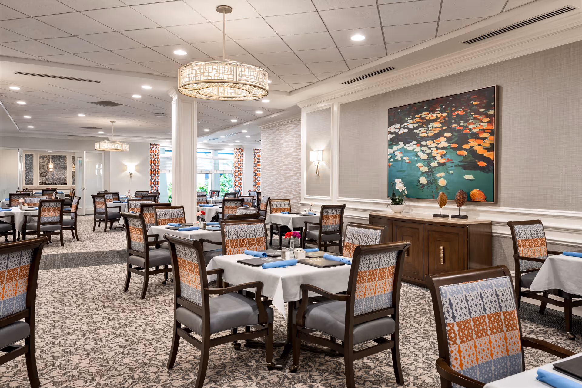 A spacious and well-lit dining room with multiple tables covered in white tablecloths and set with blue napkins. The chairs have patterned upholstery in shades of orange, gray, and white. The room features decorative wall paneling, a large colorful painting of water lilies, modern light fixtures, and patterned carpet. Large windows with patterned curtains allow natural light to enter the space.