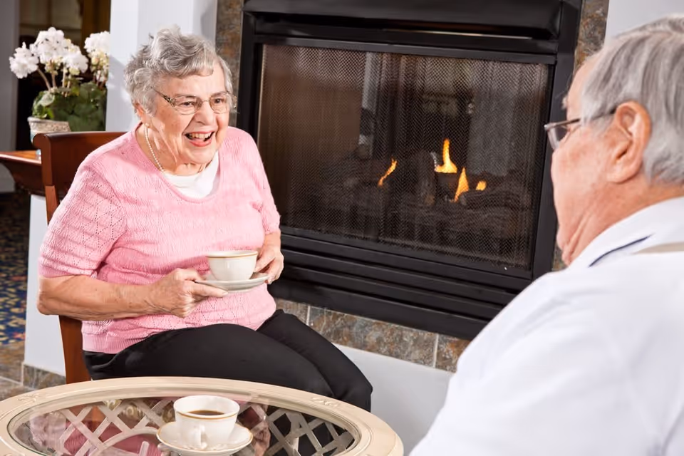 An elderly woman wearing a pink sweater and glasses is sitting on a chair, holding a cup and saucer, smiling and talking to an elderly man sitting across from her. They are seated near a lit fireplace with a glass coffee table in front of them holding another cup of coffee. There is a potted plant with white flowers in the background.