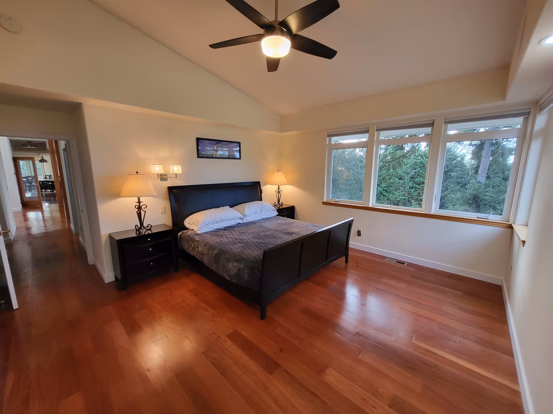 A spacious bedroom with wooden flooring, a large bed with a dark frame and gray bedding, two bedside tables each with a lamp, a ceiling fan with a light, and a row of large windows showing a view of trees outside.