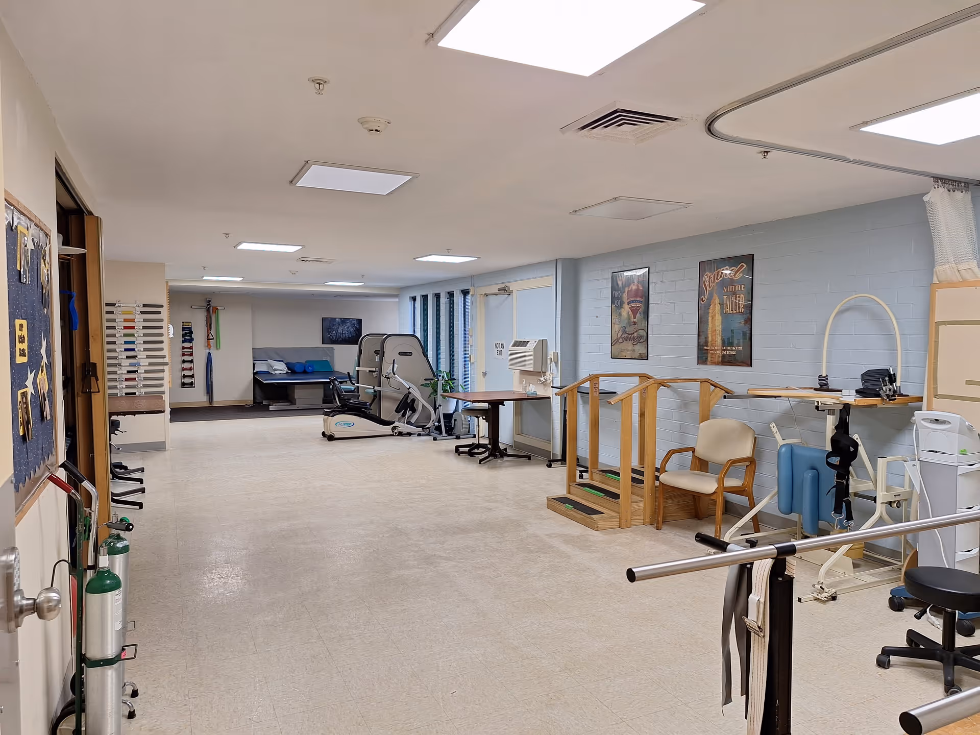 A spacious rehabilitation or physical therapy room in a senior living facility with exercise equipment, parallel bars for walking practice, chairs, and posters on the wall. The room has bright overhead lighting and a clean, organized appearance.